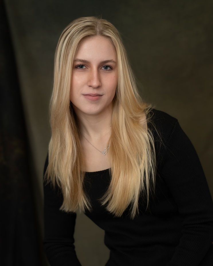 Young blonde woman in black top poses for professional portrait against dark studio background.