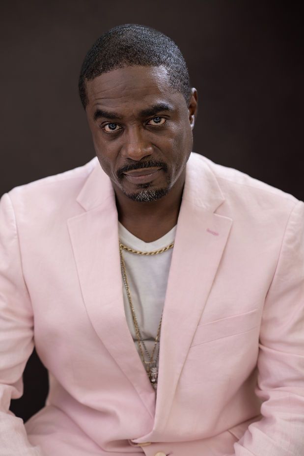 A confident man in a pink blazer and gold chain necklace poses against a dark background.