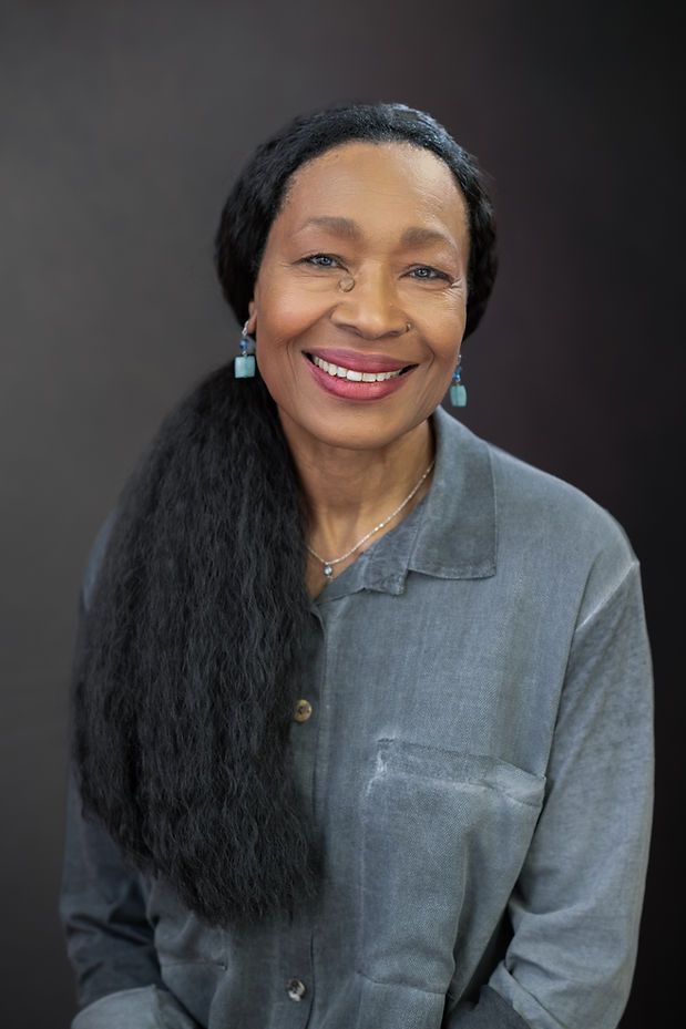 Smiling older Black woman with long black hair, teal earrings, and a gray button-up shirt posed against a dark background.