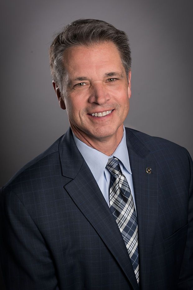 Professional headshot of a smiling middle-aged man in a navy suit and plaid tie against a grey background.