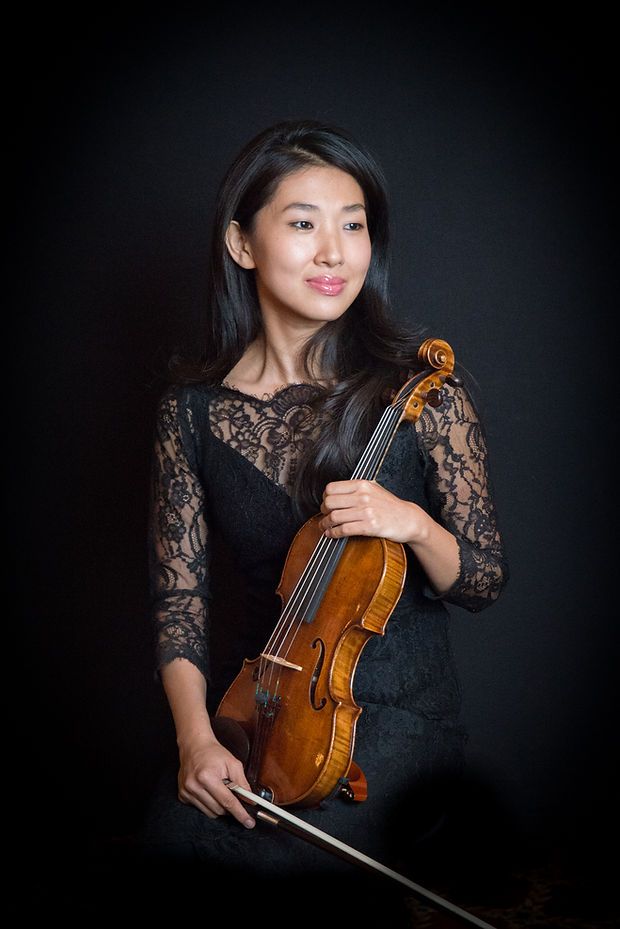 A female violinist in a black lace dress holds her violin and bow against a dark background.