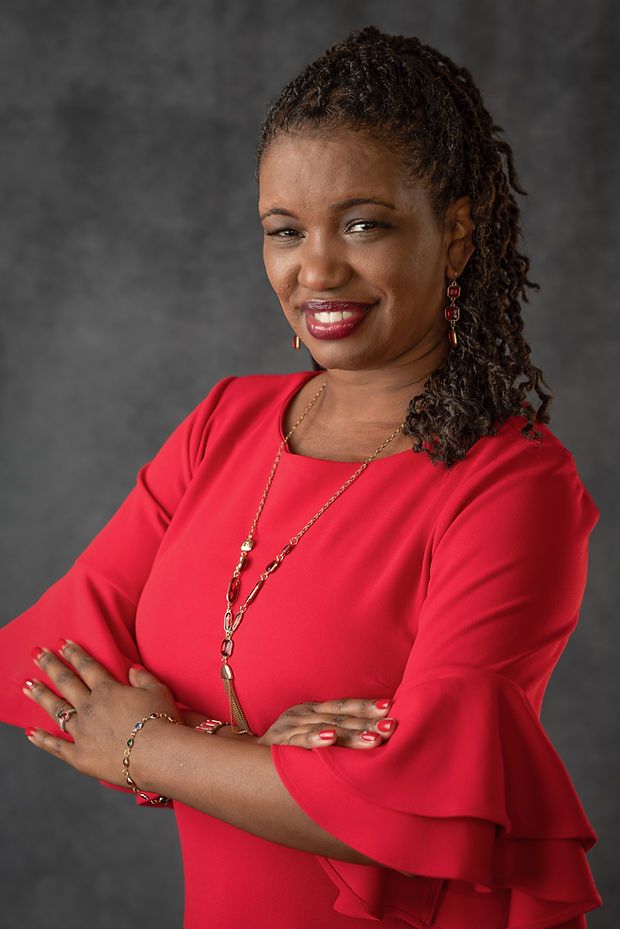 Woman in red dress with gold jewelry smiles confidently with arms crossed against gray studio background.
