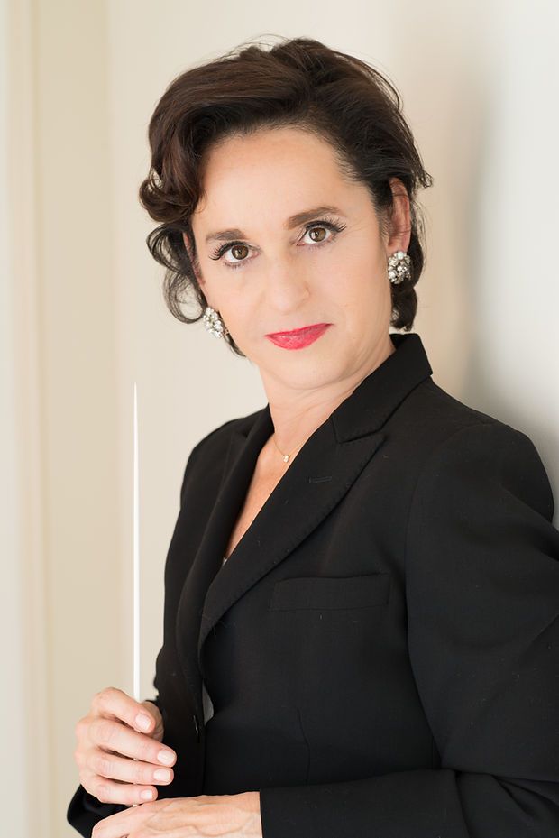 A female orchestra conductor in a black blazer holds a baton, wearing elegant earrings and red lipstick.