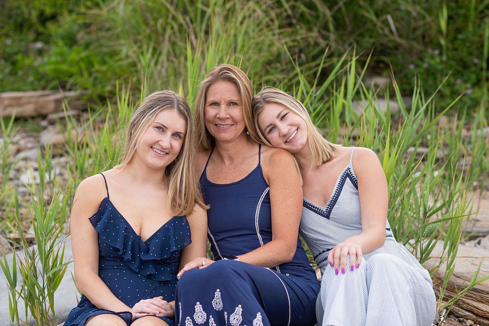 Three smiling women in blue dresses posing together outdoors near tall grass.