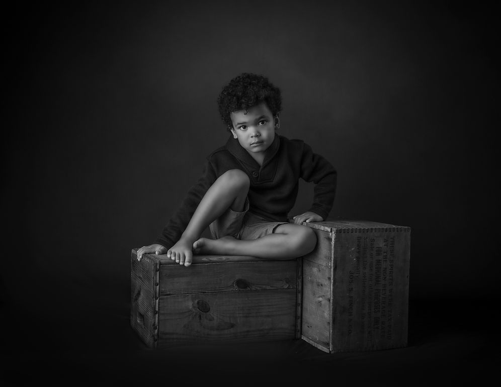 Black and white portrait of a young boy sitting on a wooden crate in a studio setting.