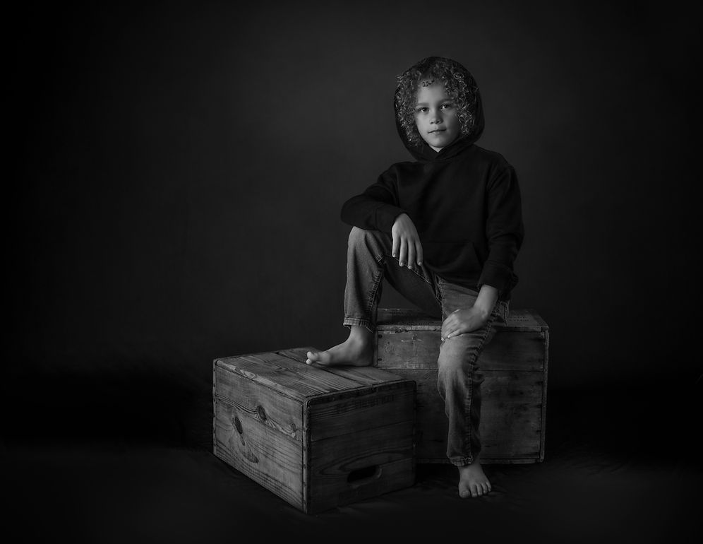 Black and white portrait of a barefoot child sitting on wooden crates in dark clothing.