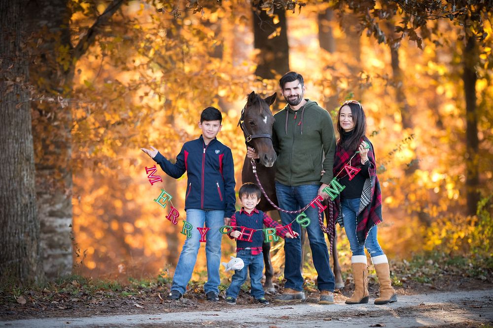 Family of five posing among vibrant autumn foliage holding a colorful Christmas banner outdoors.