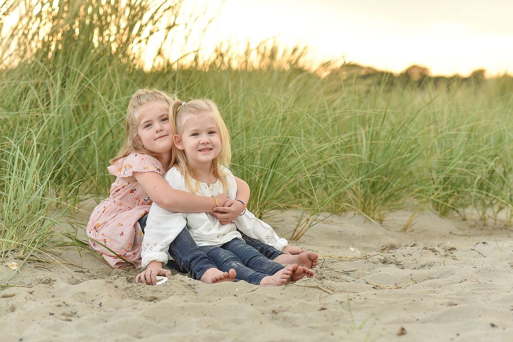 Two young sisters sitting together on sandy beach dunes hugging in golden evening light.