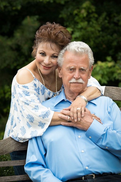 Older couple posing outdoors on a bench surrounded by lush green foliage in a warm portrait.