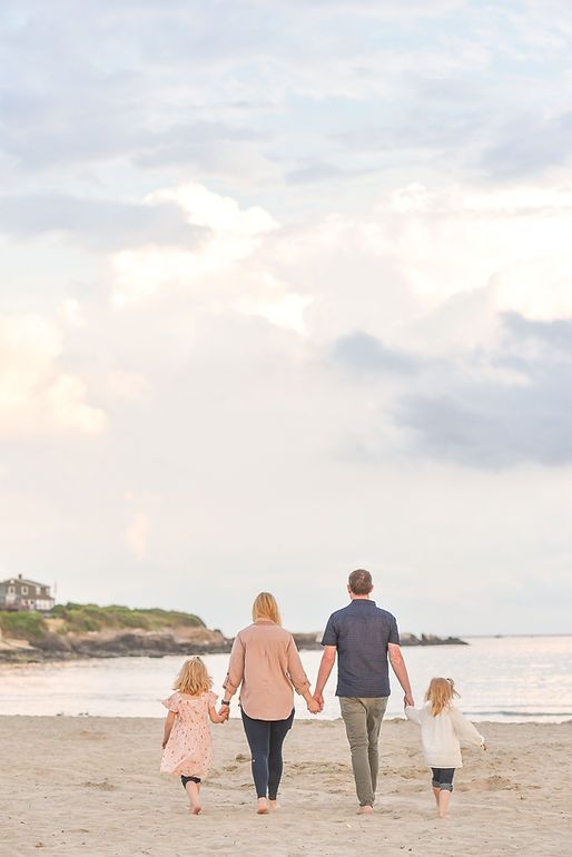Family of four walking hand in hand along a sandy beach at sunset with cloudy sky.