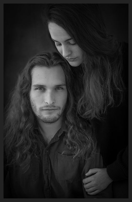 Black and white portrait of a long-haired couple posing together in dark clothing.