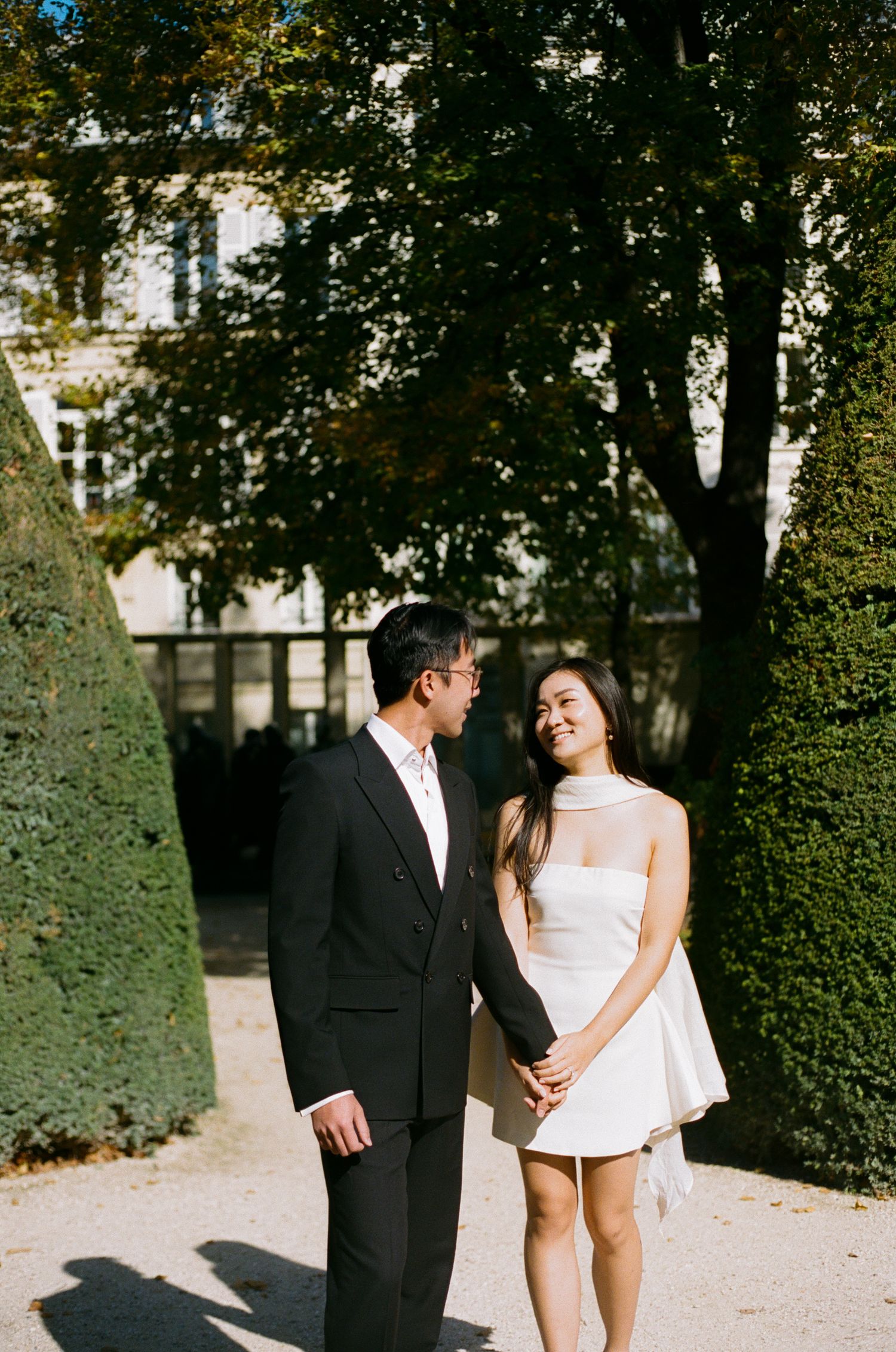 A couple shares romantic moments walking along a tree-lined path in a garden setting.