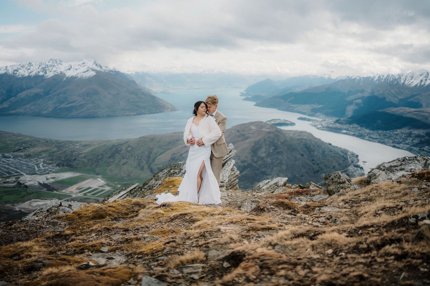 Couple in wedding attire embracing on a mountain summit overlooking snow-capped peaks and misty valleys.