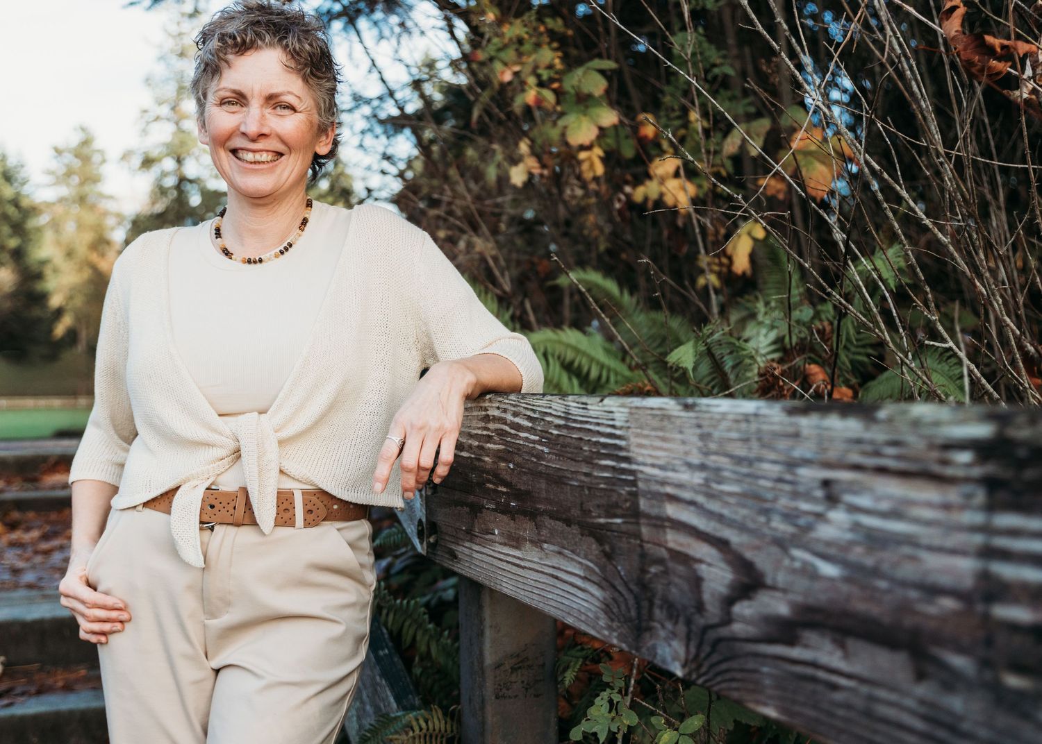 Woman in gray sweater and dark jeans poses against autumn forest backdrop near rocky shoreline.