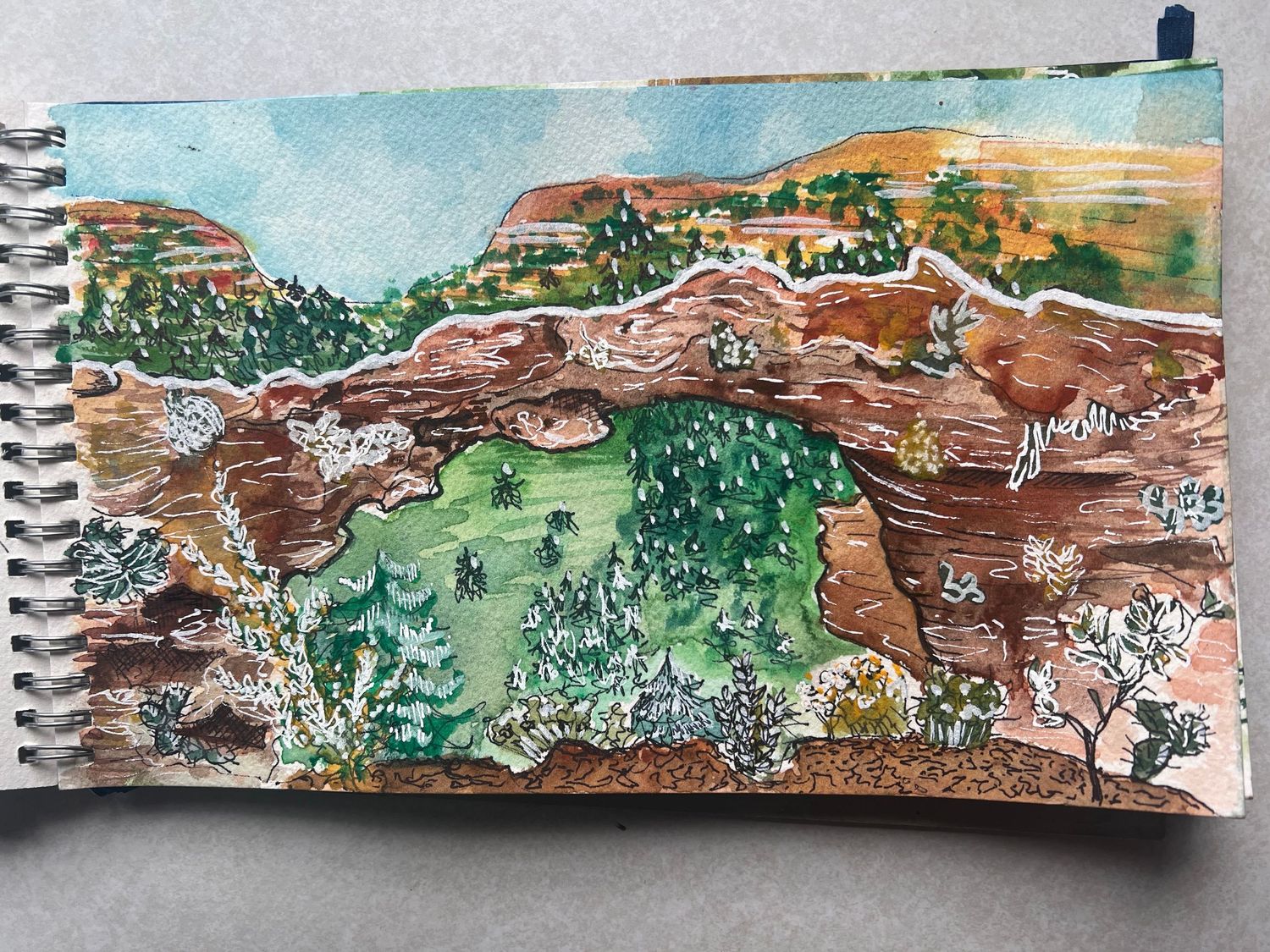 A watercolor sketch in a spiral notebook shows red rock formations and green vegetation in a desert landscape.