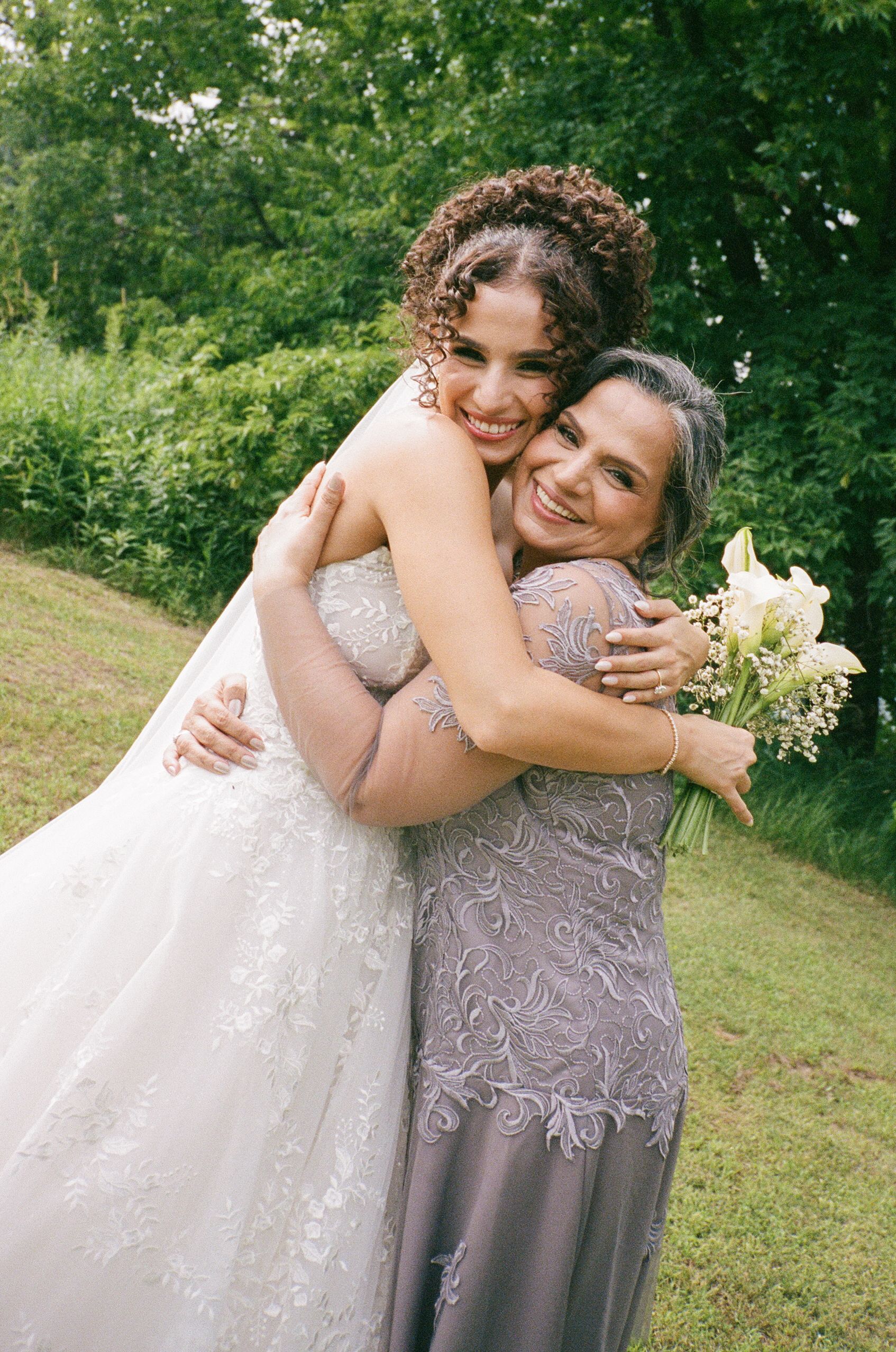Two people in formal attire share a warm embrace on a grassy lawn with green trees in the background.
