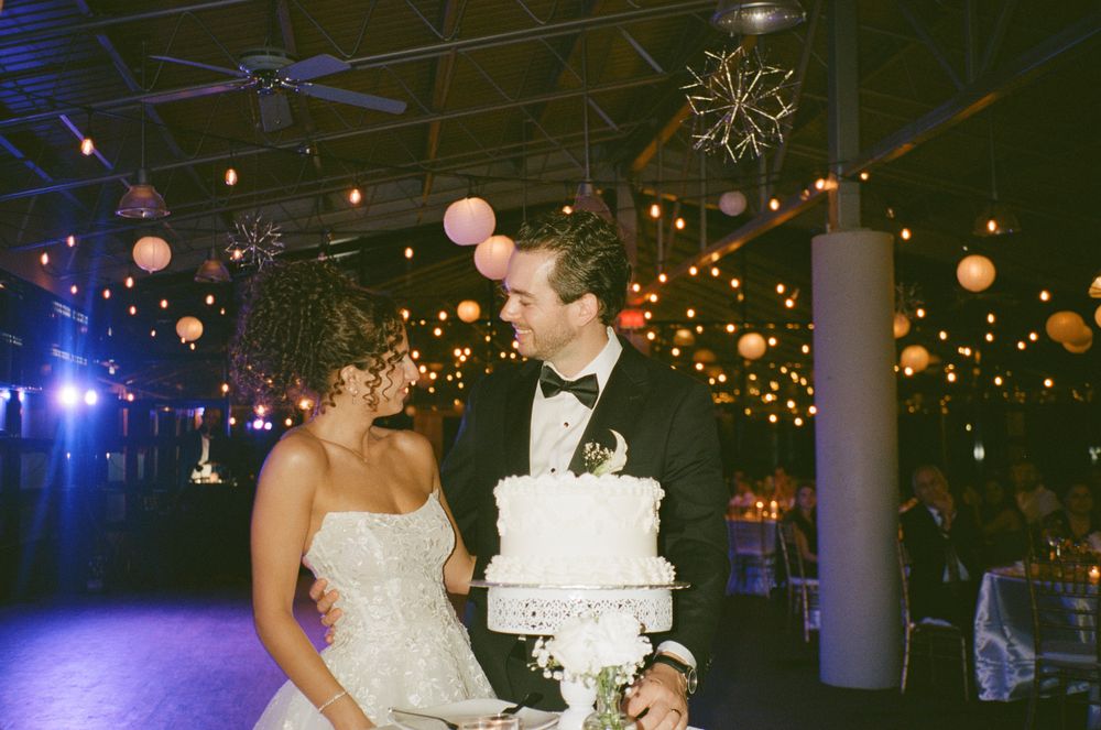 A couple cuts their elegant white wedding cake under glowing string lights and paper lanterns at an evening reception.