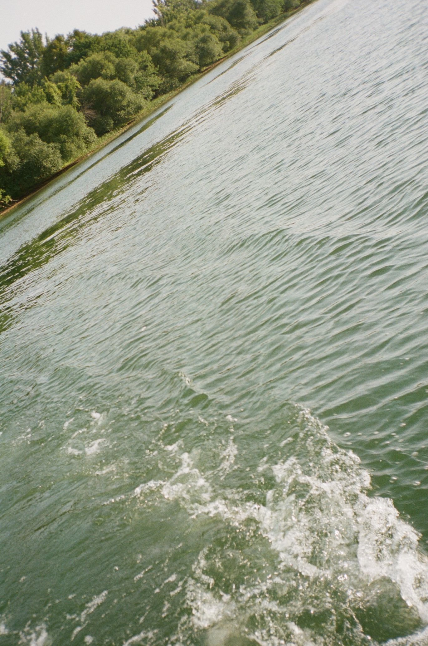 Rippling green water surface with splashing waves near a tree-lined shoreline on a cloudy day.