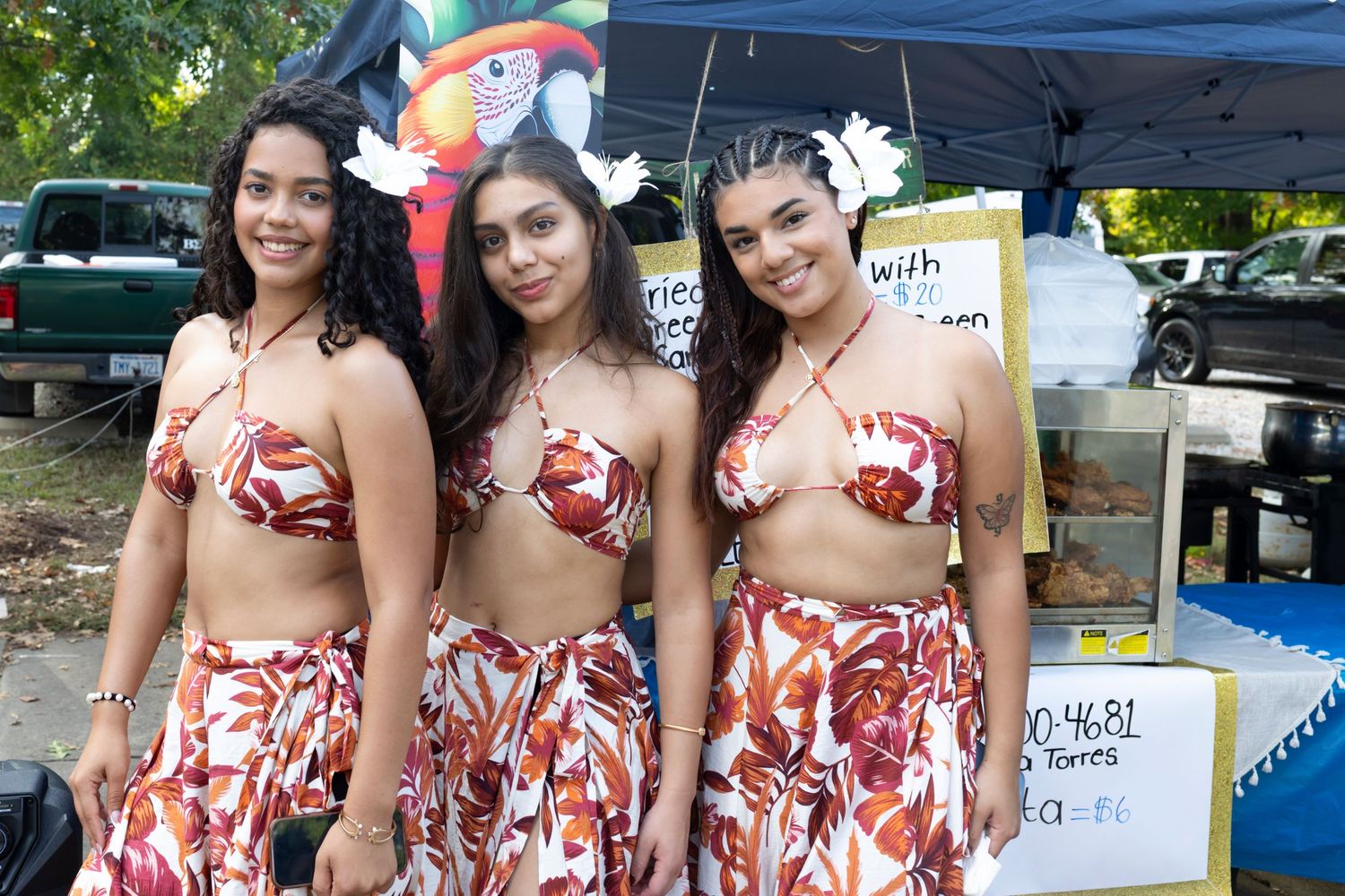 Three dancers in matching red floral print outfits pose together at an outdoor event under a blue tent.