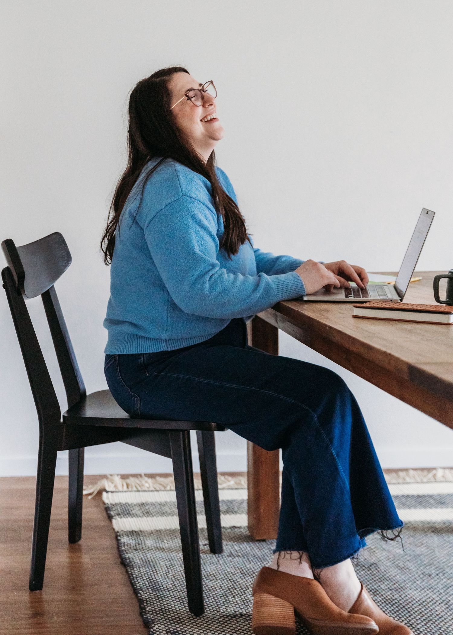 A person in a blue sweater and jeans works at a wooden desk with a laptop in a minimalist home office setting.