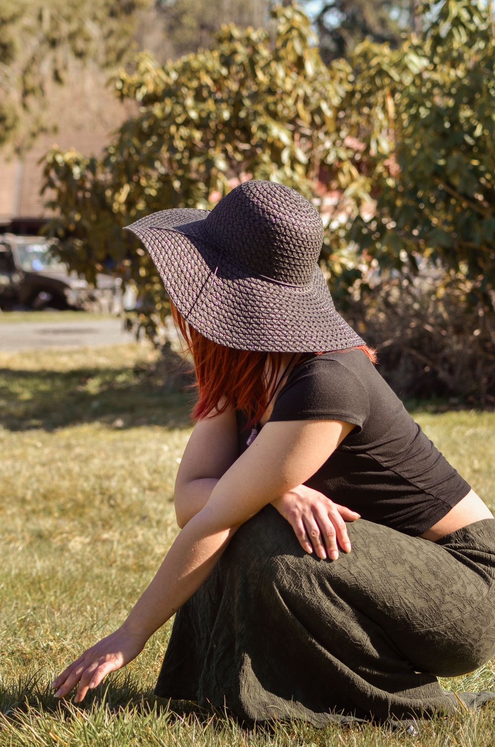 A person wearing a sun hat and dark clothing sits on grass in a park during a sunny day.