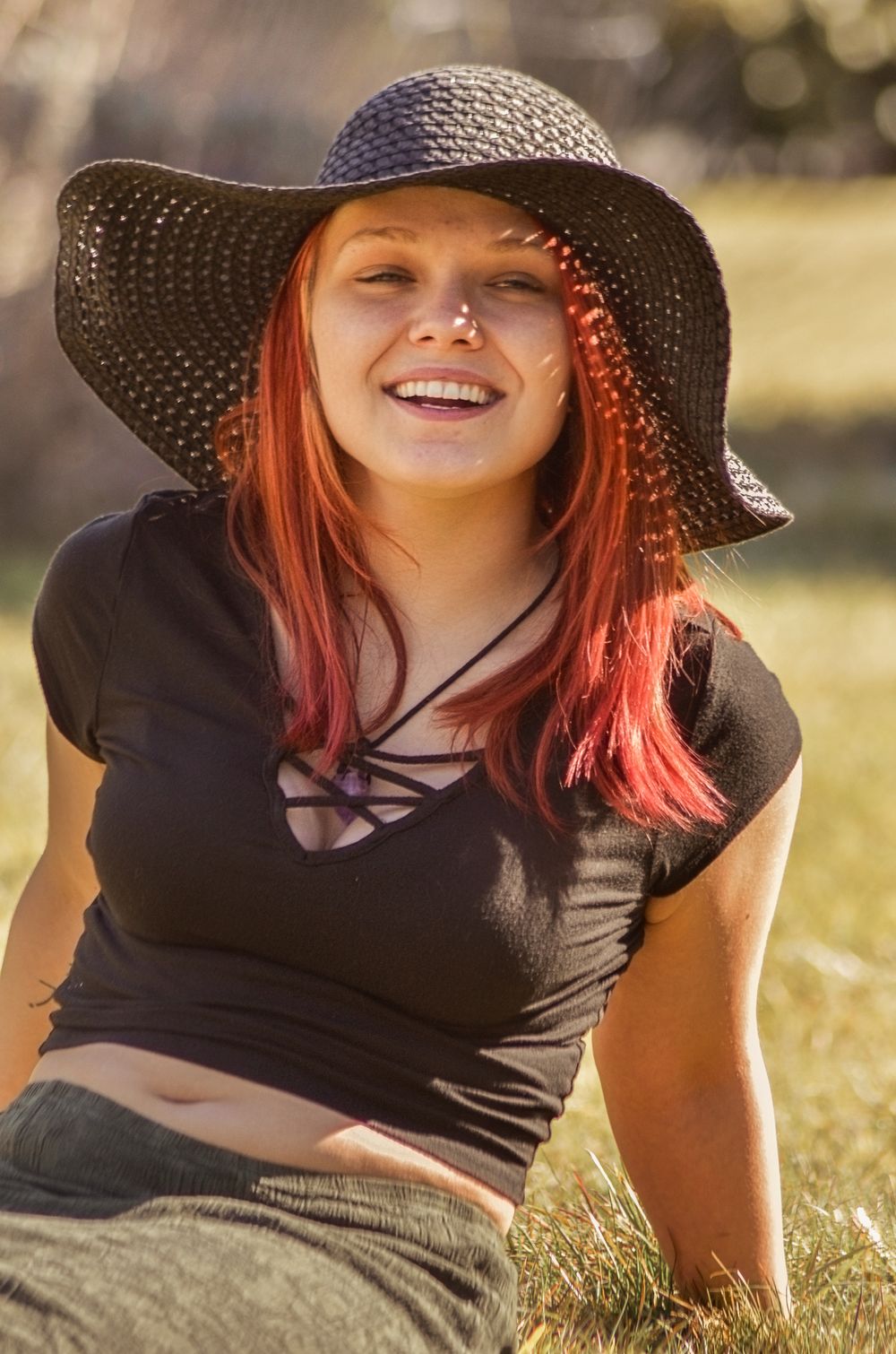 A figure in a black sun hat relaxes in golden sunlit grass with a joyful expression.