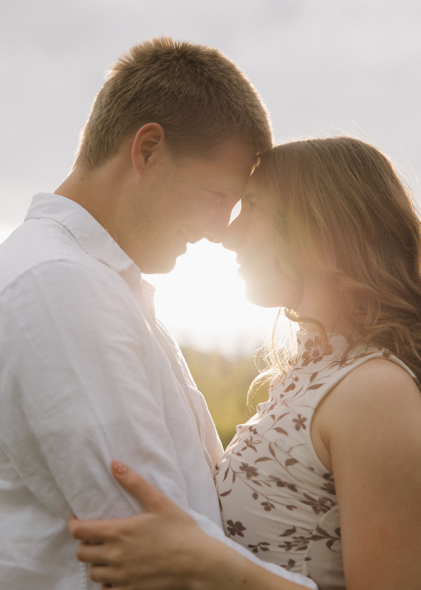 Romantic couple touching foreheads and smiling outdoors among green trees in a tender intimate moment.