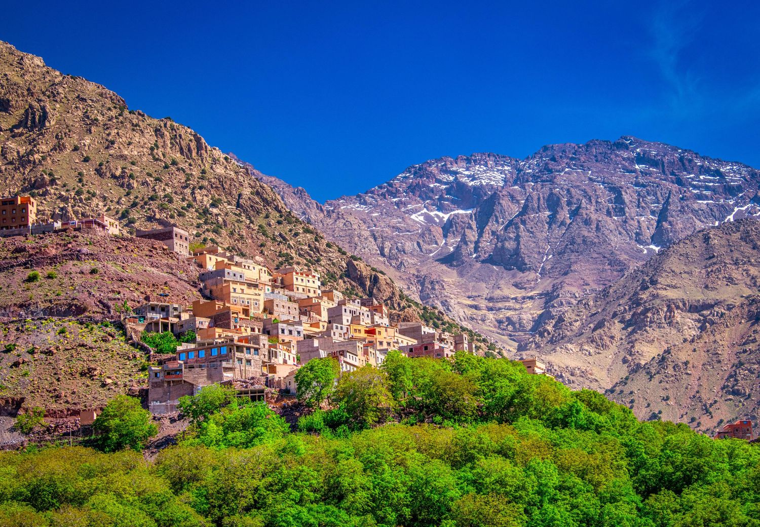 Panoramic view of traditional Moroccan mountain village nestled in Atlas Mountains with lush green vegetation below.