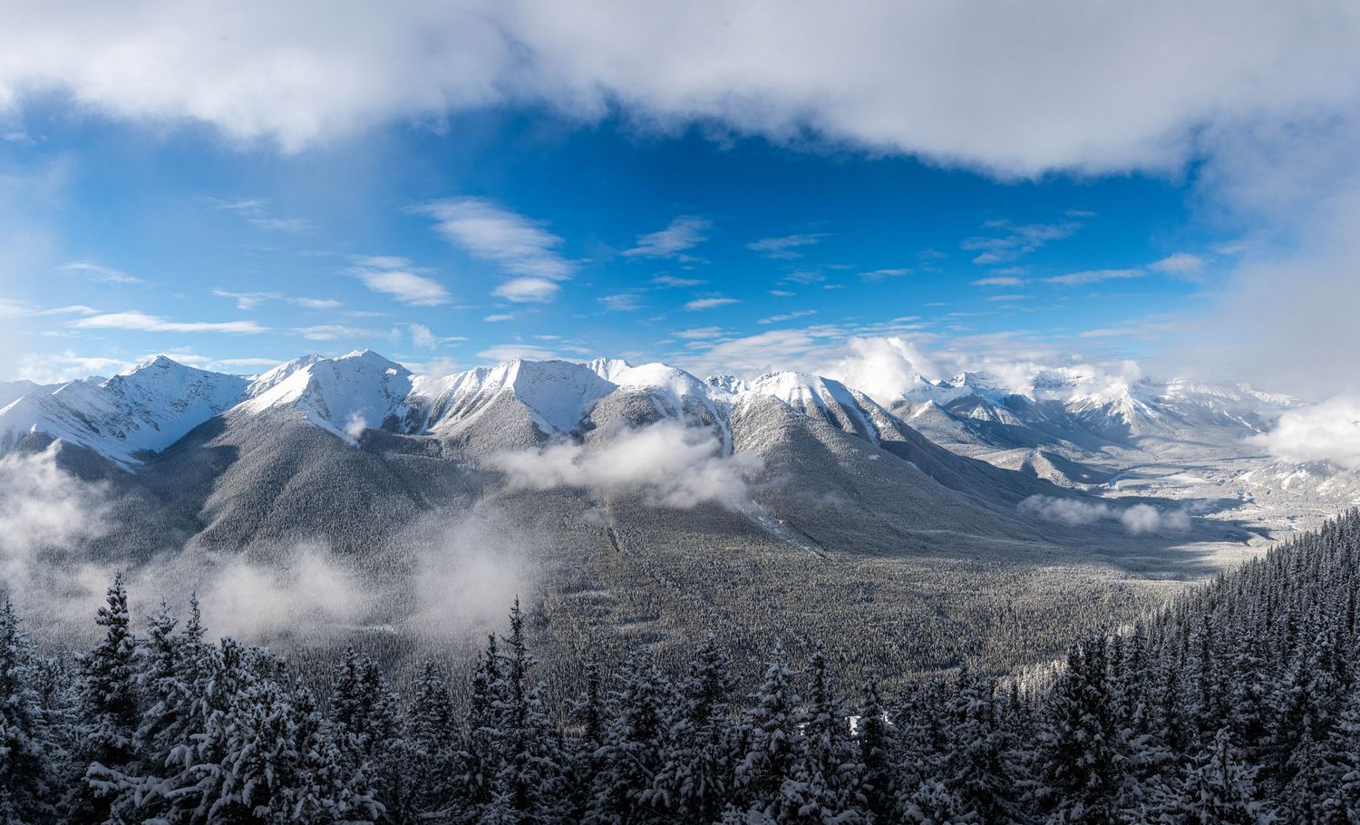 Snow-capped mountain peaks rise against a blue sky with scattered clouds and evergreen forests in the foreground.