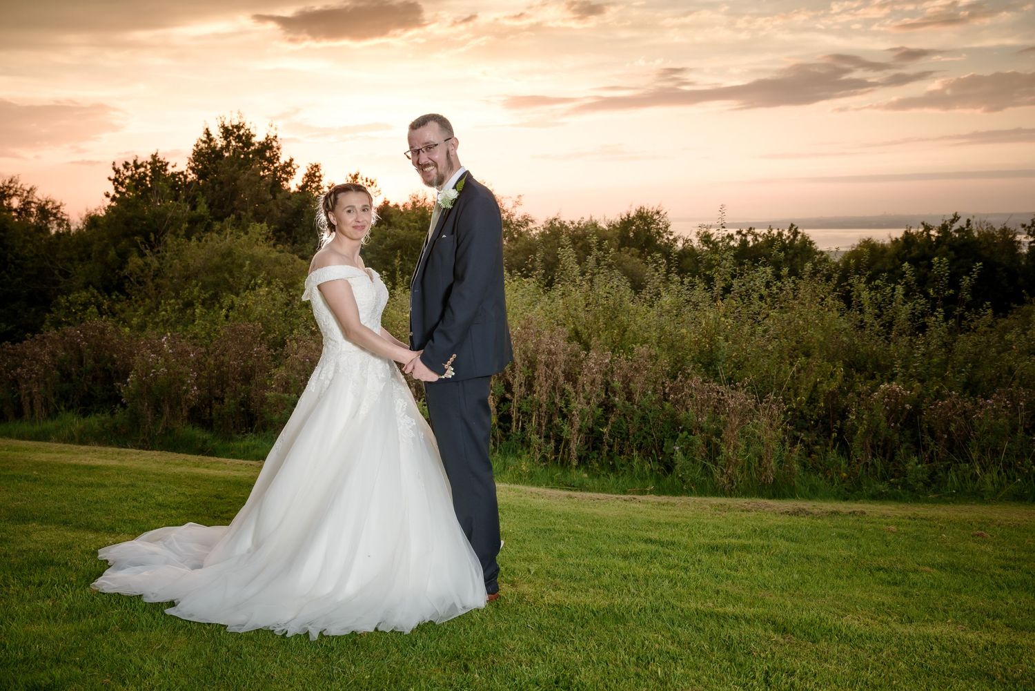 A couple in wedding attire poses together on a grassy hill at sunset with trees in the background at Forest Hills Hotel in Frodsham