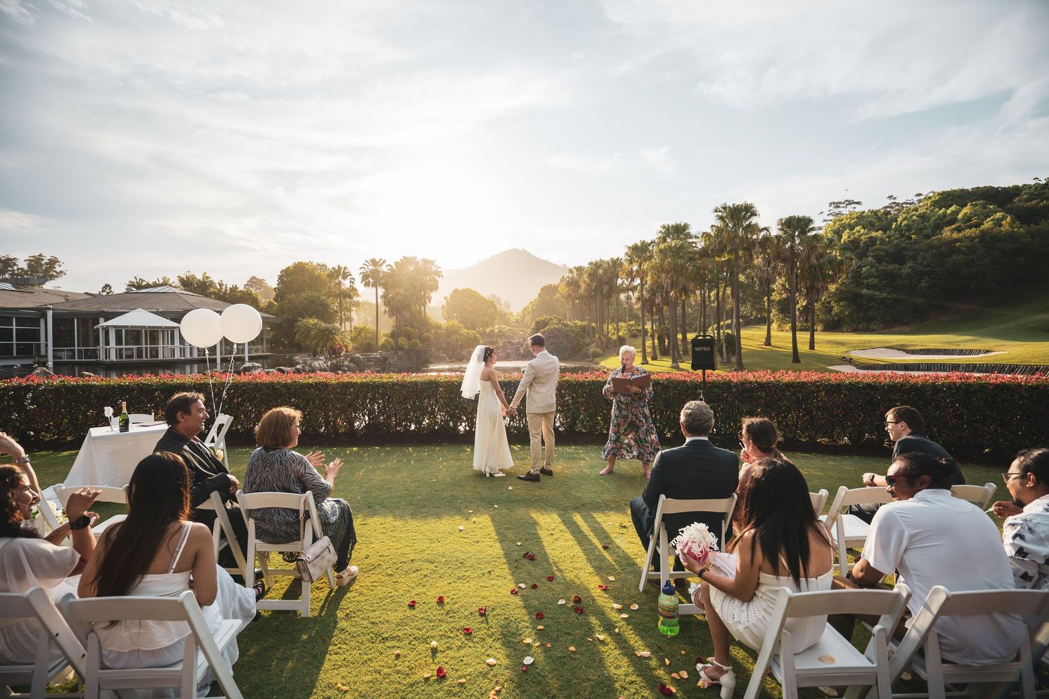 Wedding guests gather outdoors at sunset for a celebration on a grassy lawn with palm trees in the background.
