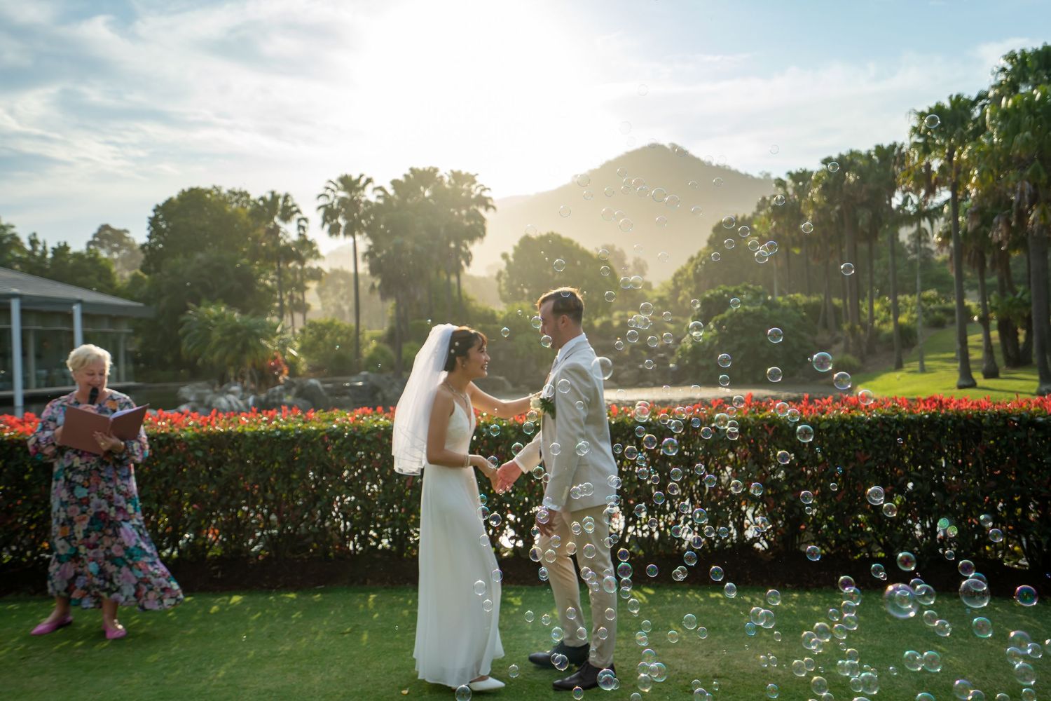 People gather in a sunlit garden surrounded by vibrant flowers and mountains during a scenic outdoor event.