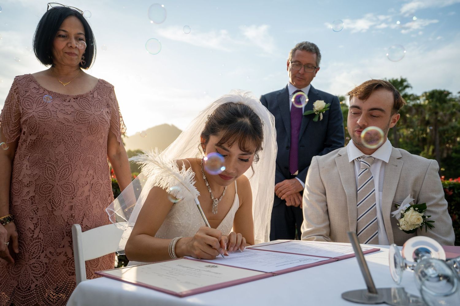 Wedding guests sign the marriage register at an outdoor ceremony with trees in the background.
