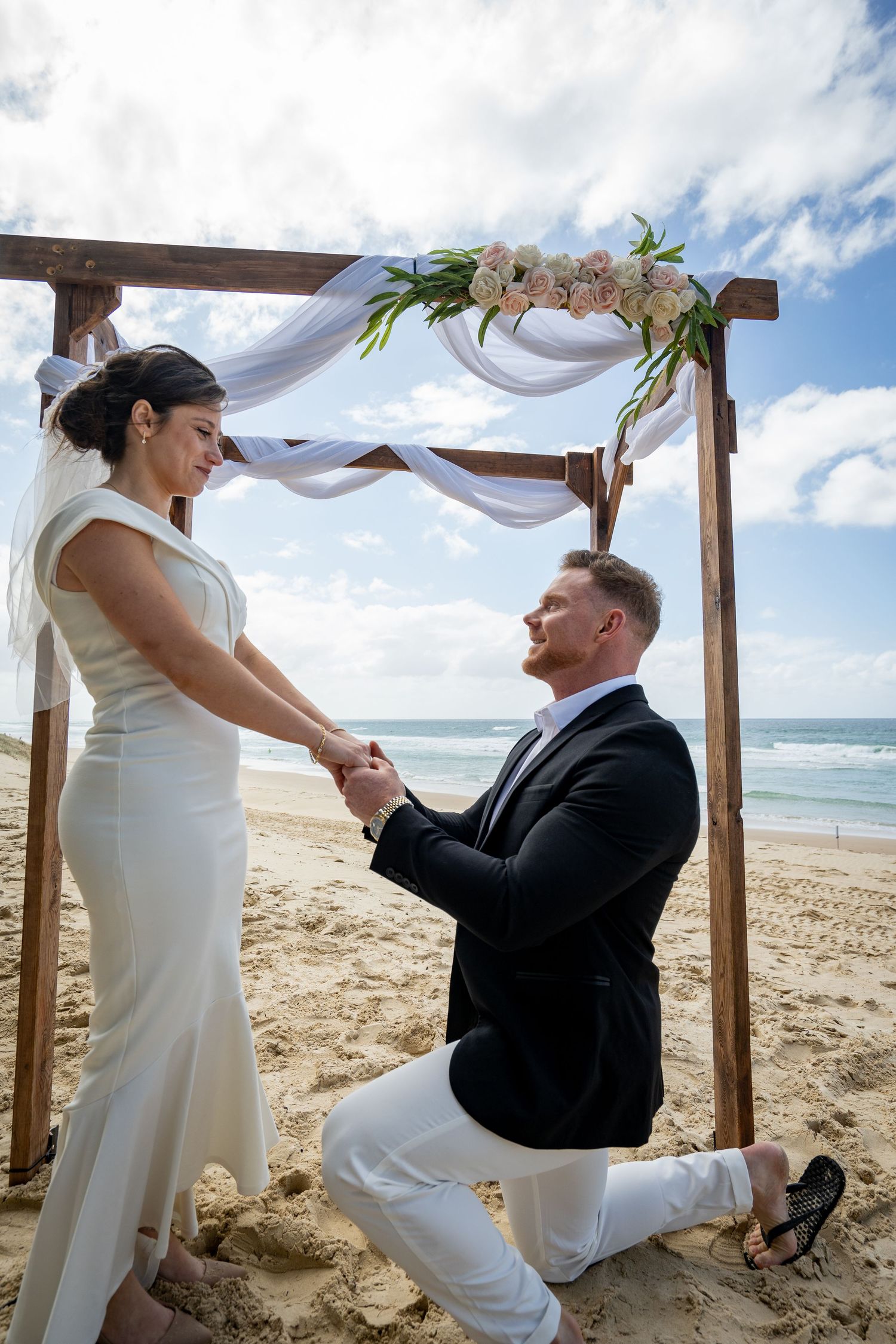 A romantic beach wedding proposal scene with wooden arch and flowing white fabric against ocean backdrop.