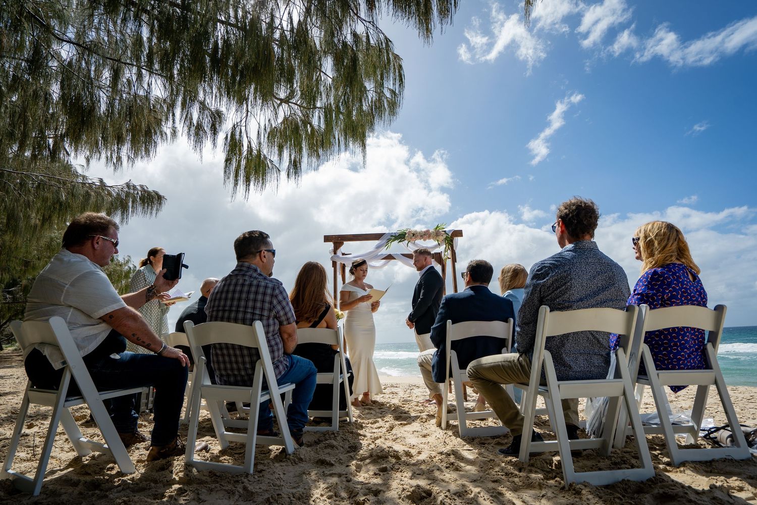 People sitting in white chairs on a beach watching the sunset through palm trees and Spanish moss.