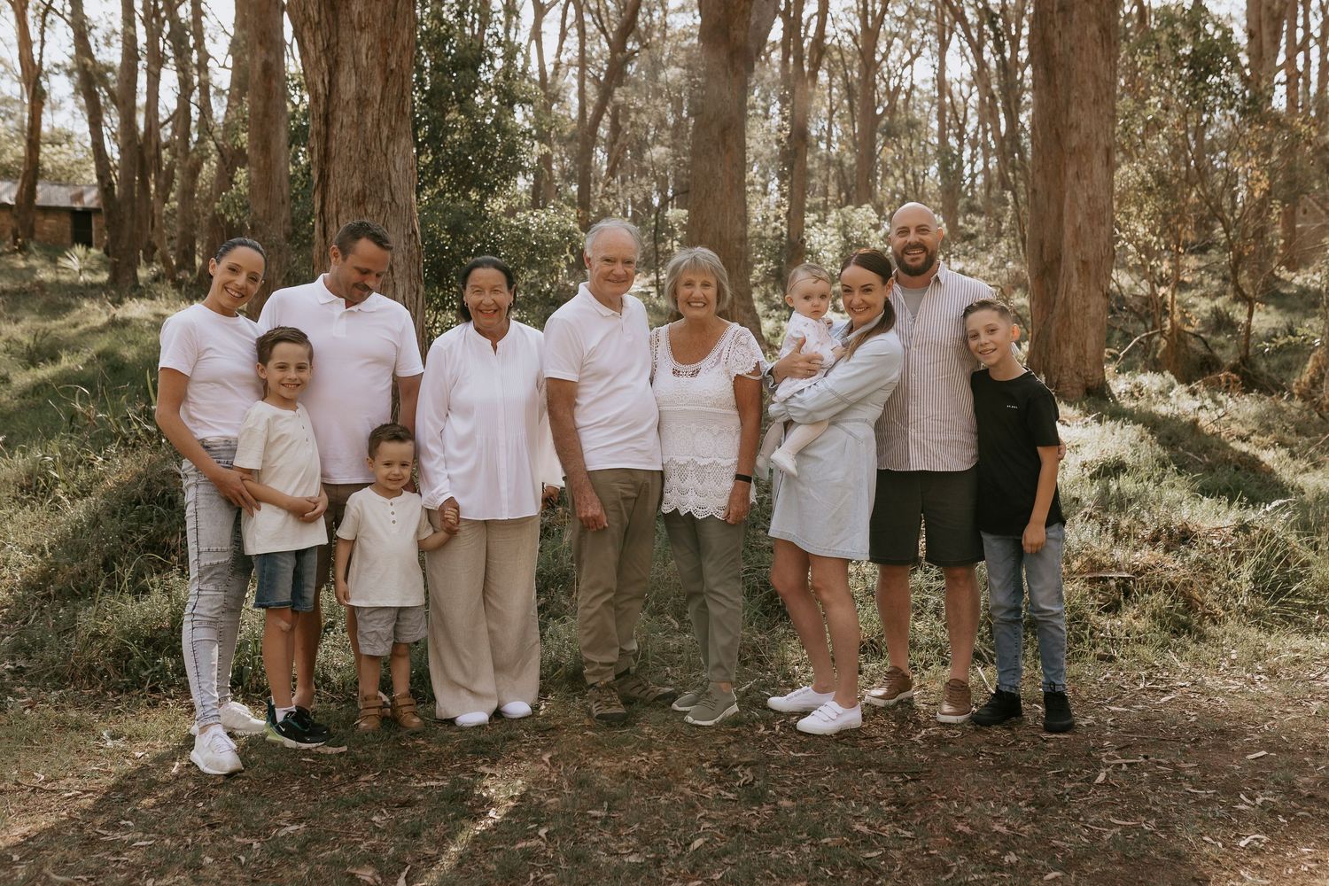 A group wearing coordinated white outfits poses together in a wooded outdoor setting for family photos.