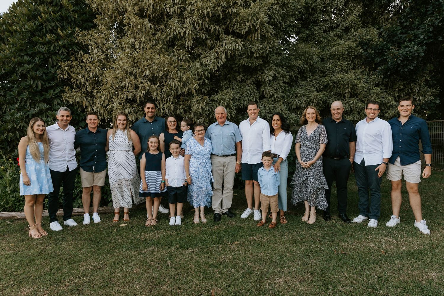 A group of people in casual summer attire stand together outdoors for a family photo session in a grassy area.