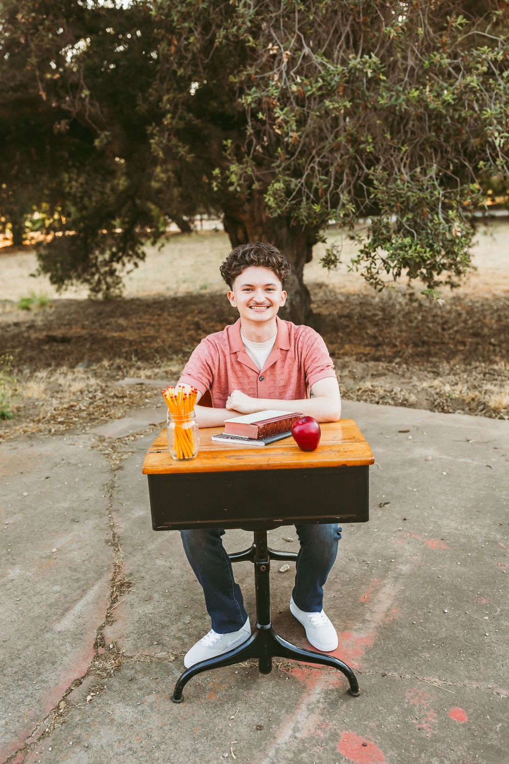 Student in pink shirt sits at wooden table with orange pencils and red cup in outdoor setting.