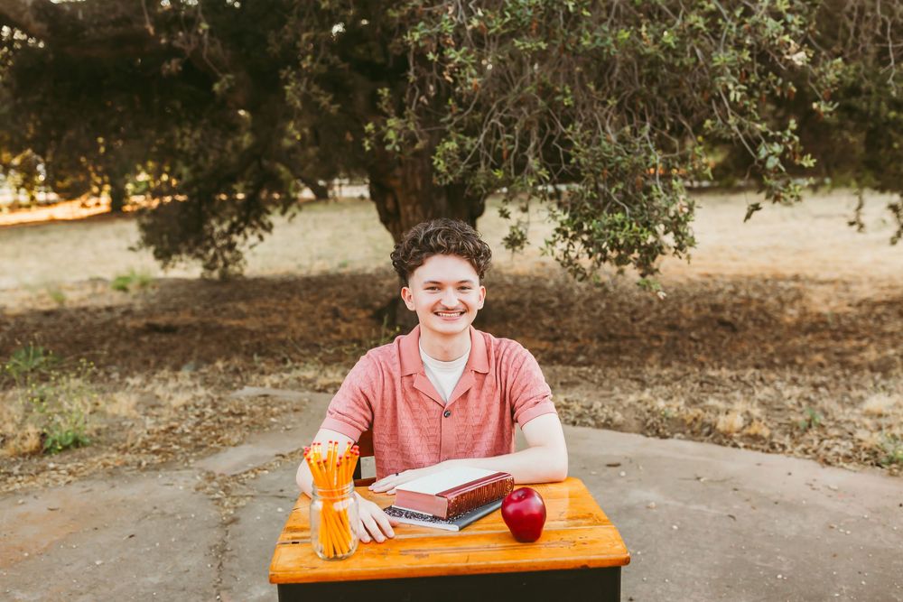 A student sits at an outdoor desk working on homework with a red apple and orange pencil case.