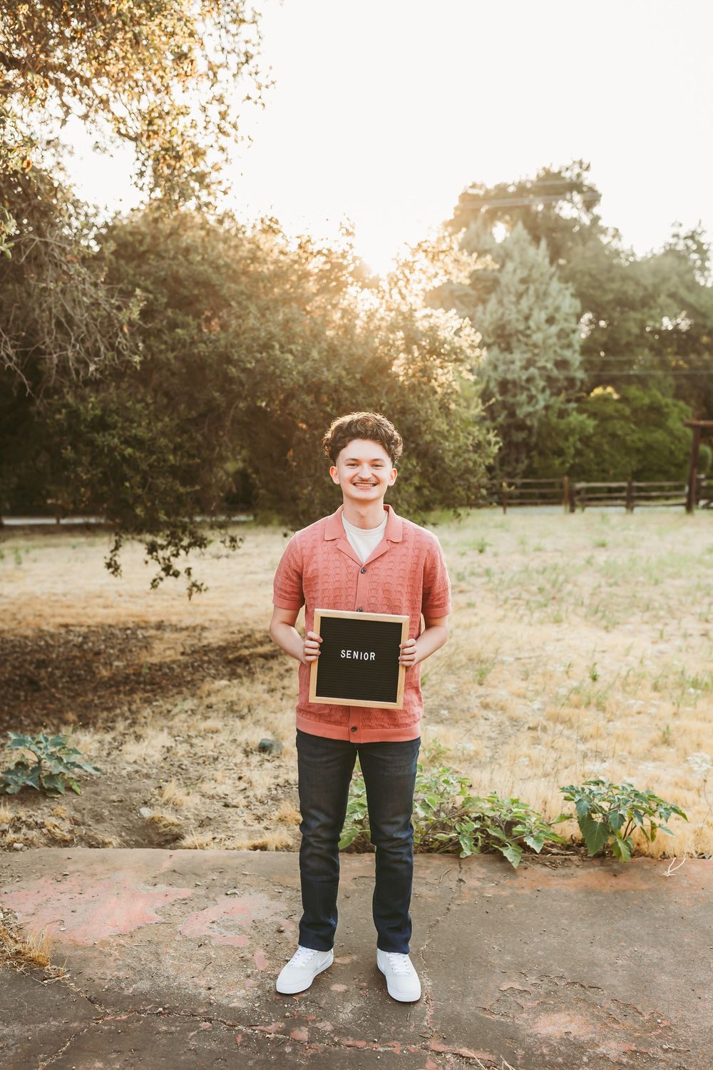 Person wearing coral shirt and jeans stands in a park setting with a chalkboard.