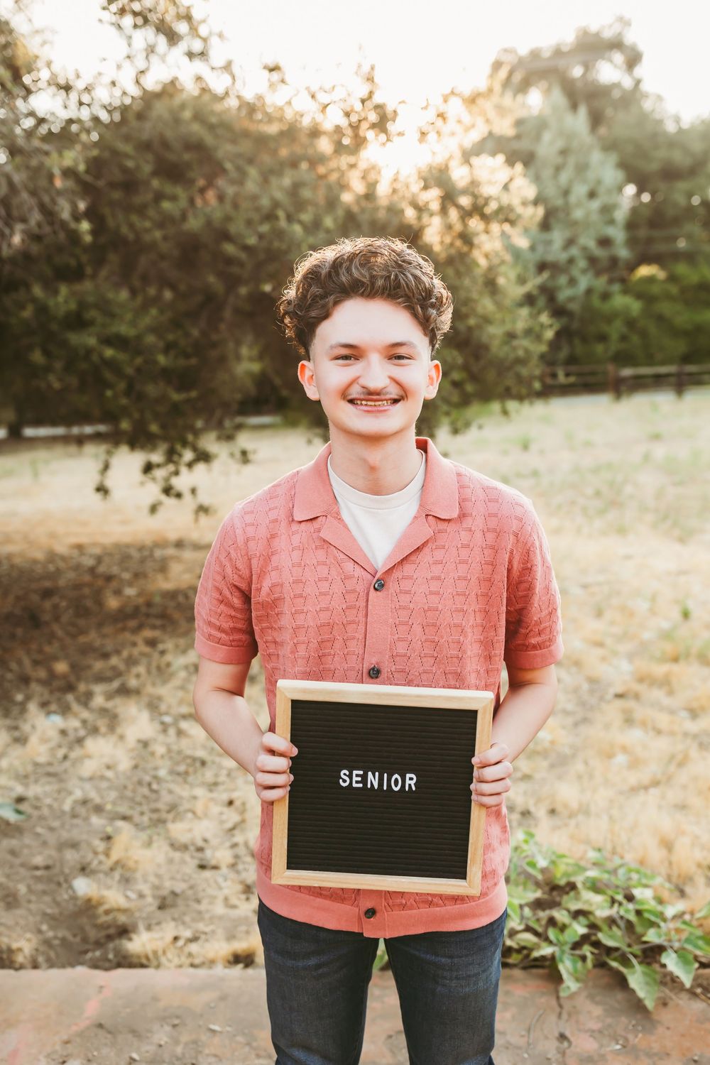Student in a coral button-down shirt holding a letter board with 'SENIOR' text in an outdoor setting.