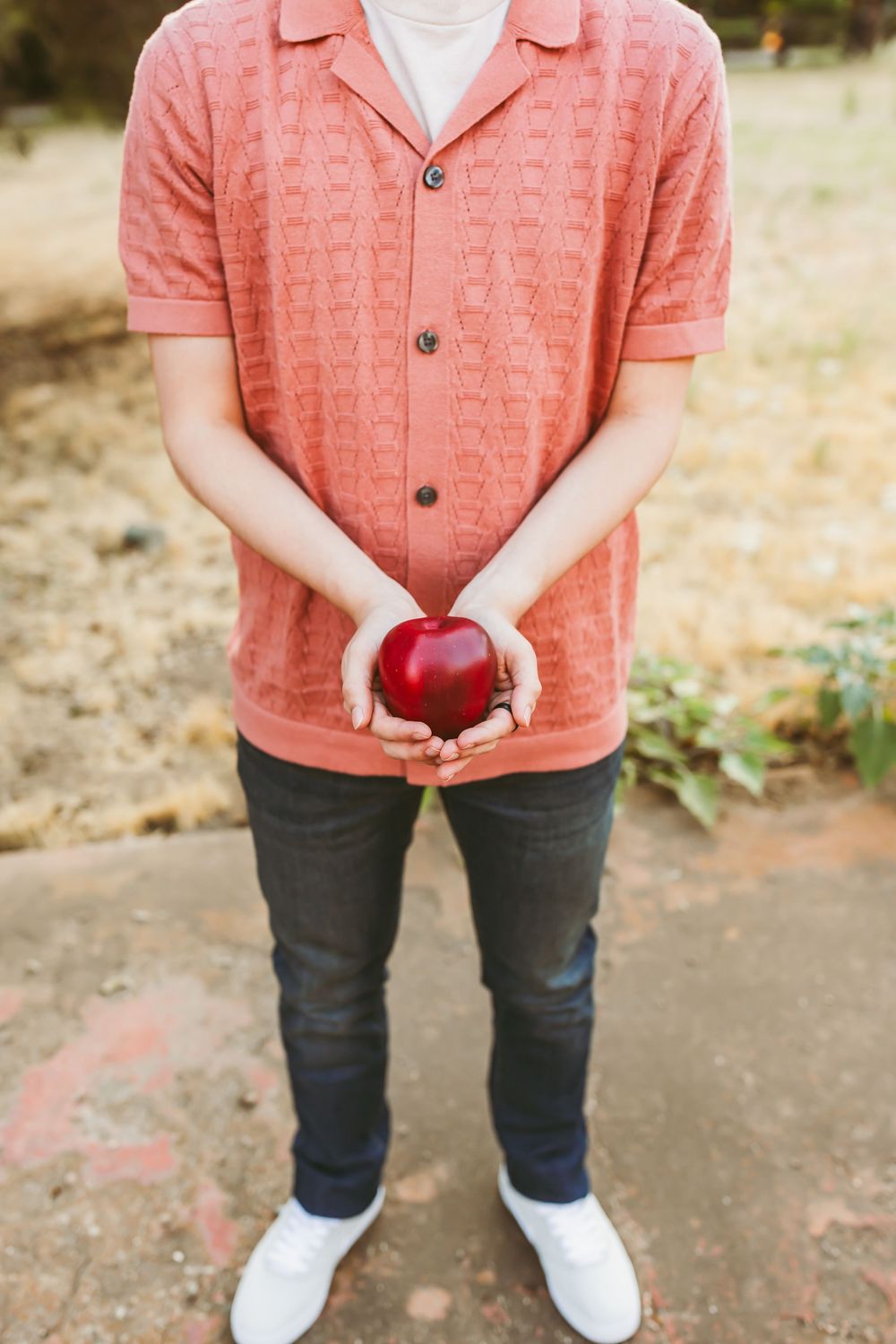 Person in a coral knit polo shirt and dark jeans holds a red apple against an outdoor background.