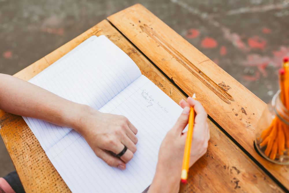 Hands write on paper at a wooden desk with orange pencil case and scattered school supplies.