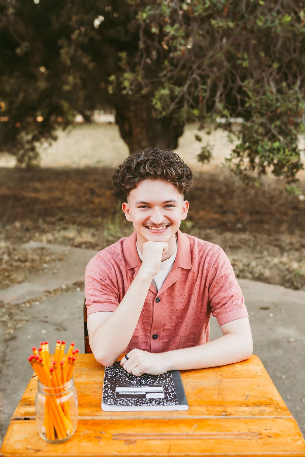Student with composition notebook sits at wooden desk with orange pencils outdoors.