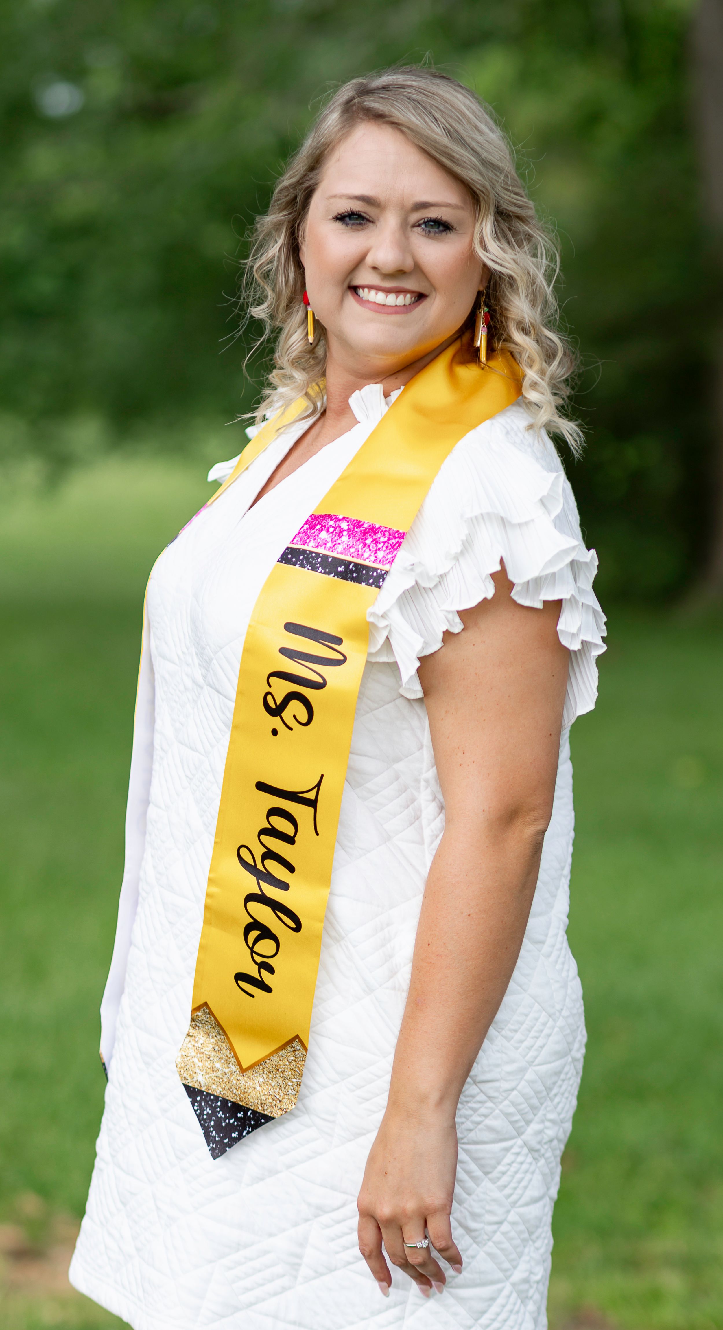 Graduate wearing a white dress and yellow sash poses for celebratory photos outdoors in a natural setting.