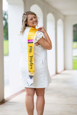 A toddler in a white dress plays with a yellow measuring tape in an outdoor covered walkway.