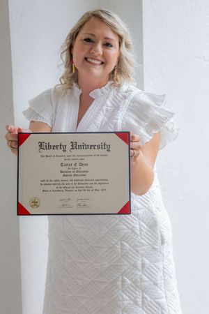 Graduate in white dress displays Liberty University diploma while posing against white background.