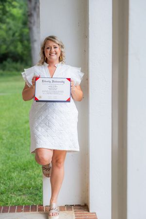 A graduate in a white dress holds up their diploma while posing between white columns outdoors.