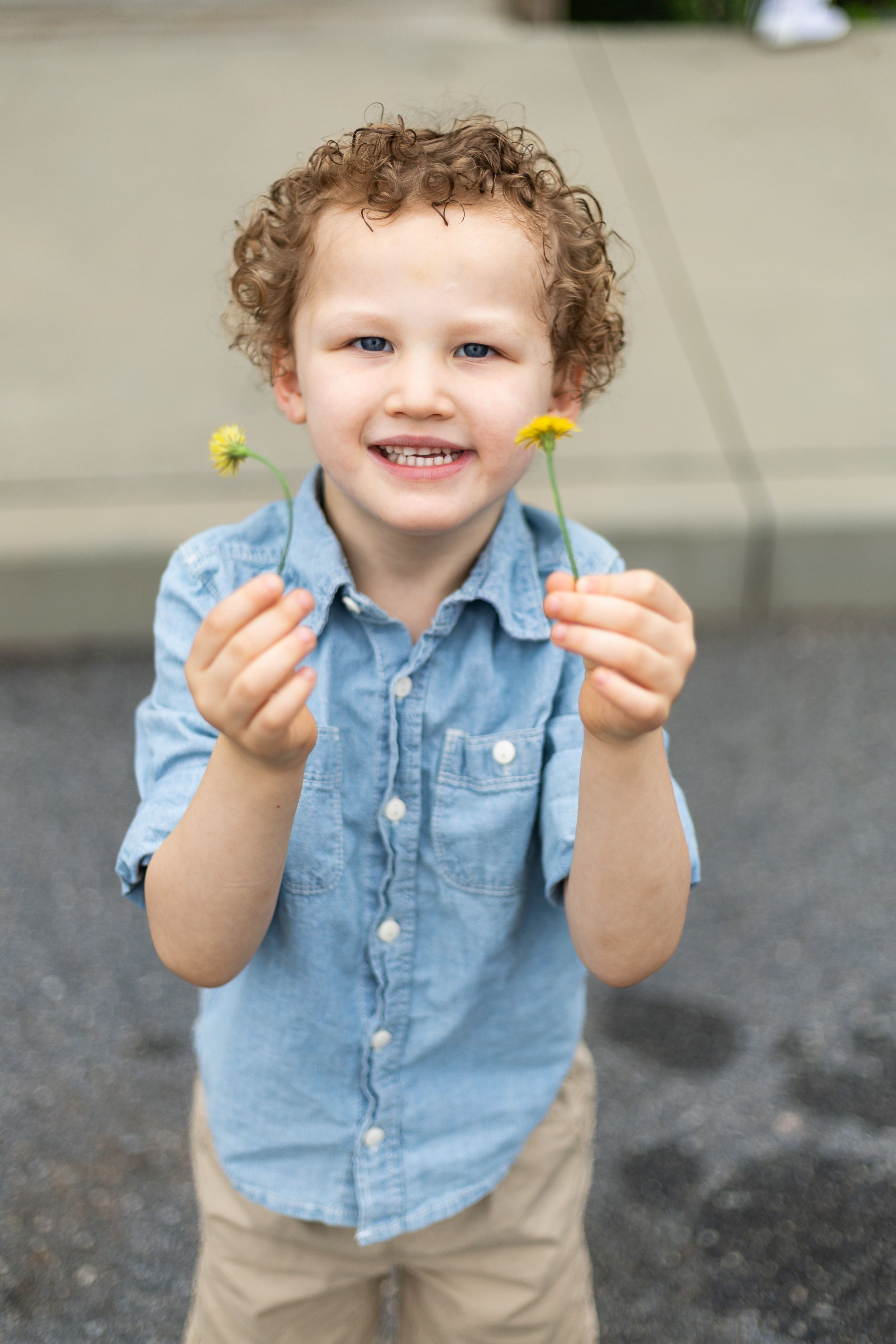 Young child in denim shirt holds yellow dandelion flowers while smiling outdoors.