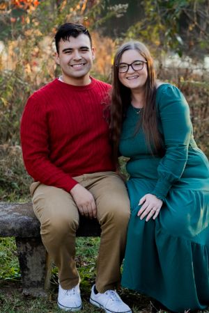 Couple in festive red and green outfits sitting together on stone wall during autumn.